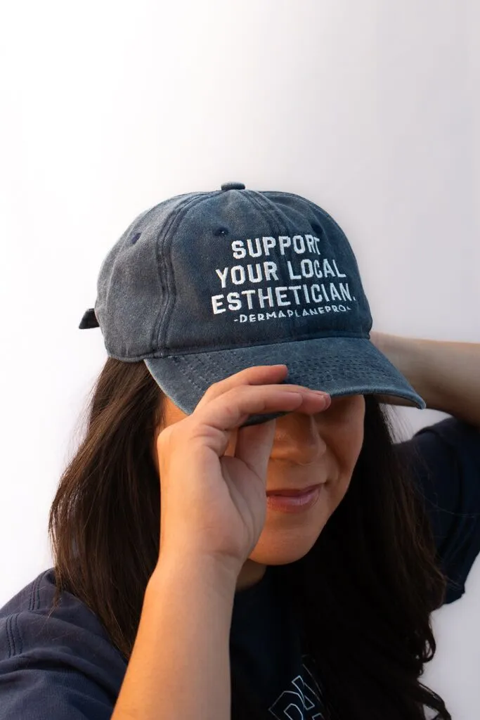 dermaplanepro-support-your-estie-hat Woman putting on a DermaplanePro distressed blue dad hat reading "Support Your Local Esthetician".