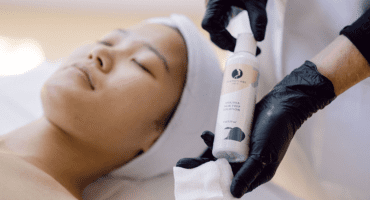 Hands wearing black latex gloves is showing the viewer a bottle of AHA/BHA Skin Prep and cotton gauze. A client is visible with her eyes closed.